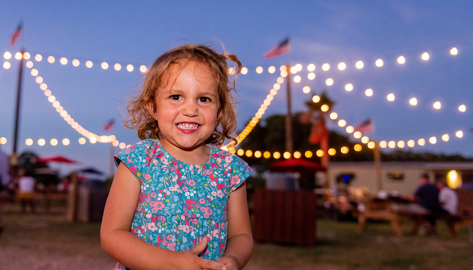 BLOG A smiling child wearing a floral dress stands outdoors at dusk with string lights and American flags in the background.