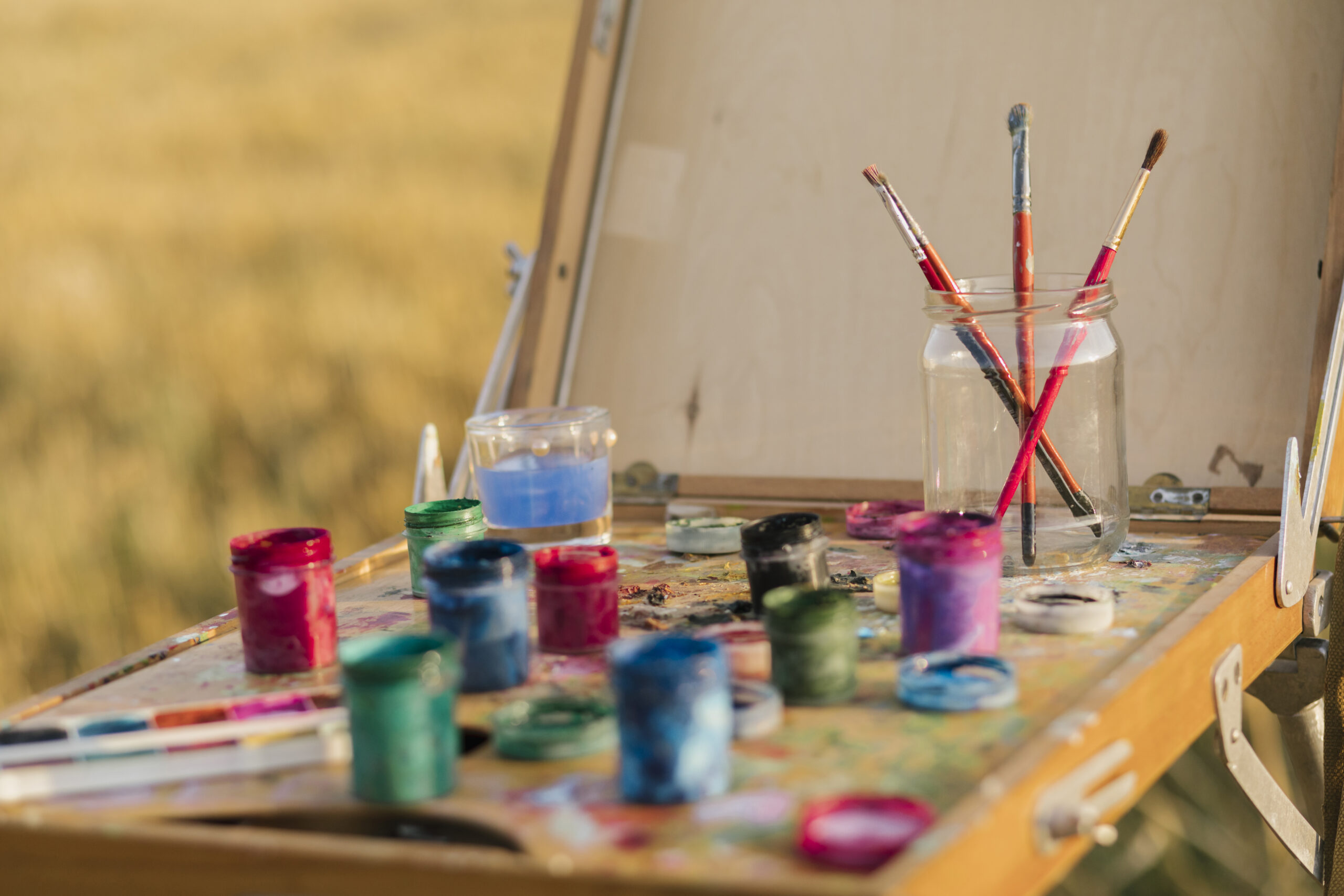 close-up-set-painting-elements Close-up of a painter's easel with an assortment of colorful paint jars and brushes in a jar, set outdoors against a blurred background of a field.